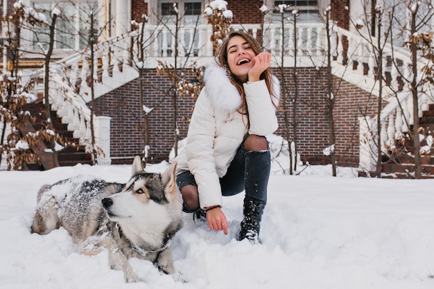 Vrolijke modieuze jonge vrouw met plezier met husky hond in de sneeuw op straat buiten. houd van huisdieren, mooie momenten, glimlachen, echte positieve emoties uitdrukken.