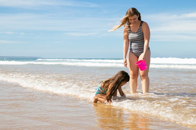 Vrolijke moeder en dochtertje staan enkel diep in zeewater en nat zand, schelpen plukken in de emmer