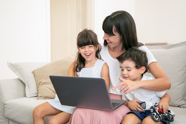 Vrolijke moeder knuffelen gelukkige kinderen terwijl ze thuis film of video kijken op laptop.