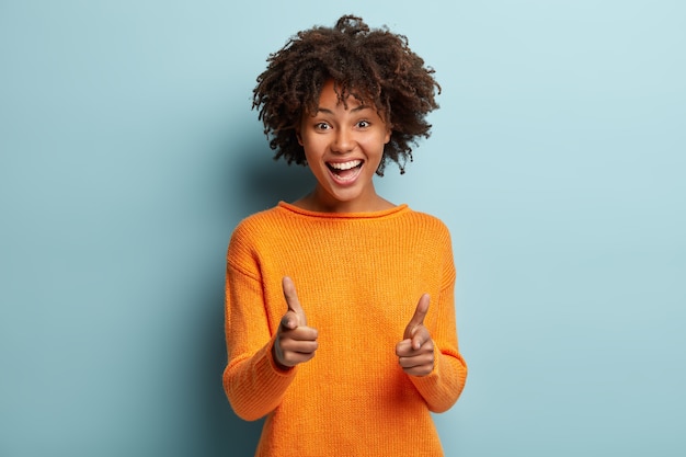 Vrolijke mooie Afro-Amerikaanse dame maakt vinger pistool gebaar op camera, keus, glimlacht breed, gekleed in oranje trui