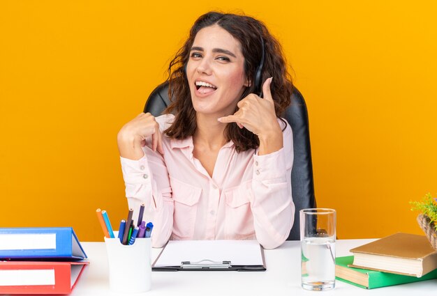 Vrolijke, mooie blanke vrouwelijke callcenter-operator op koptelefoon zittend aan bureau met kantoorhulpmiddelen gebaren bel me teken geïsoleerd op oranje muur