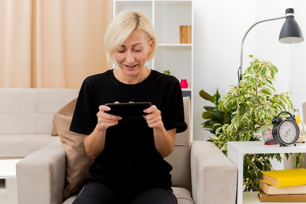 Gratis foto vrolijke mooie blonde russische vrouw zit op fauteuil te houden en telefoon te kijken