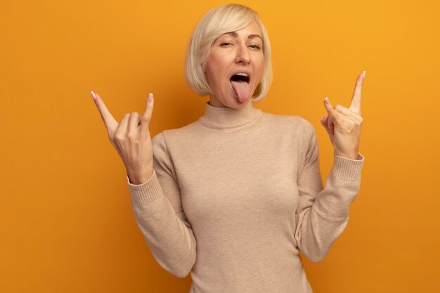 Vrolijke mooie blonde Slavische vrouw steekt tong uit en gebaren hoorns met twee handen op sinaasappel