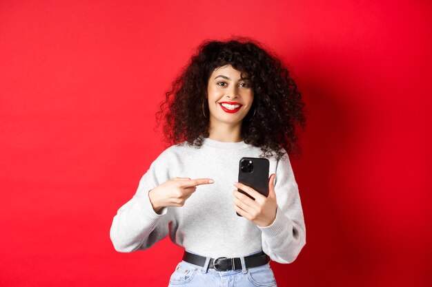 Vrolijke mooie dame met smartphone die naar haar telefoon wijst, tevreden glimlacht, een app aanbeveelt, staande tegen een rode achtergrond