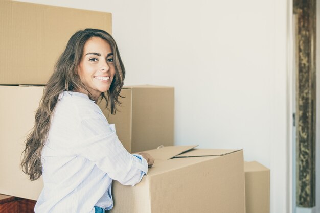 Vrolijke mooie jonge Spaanse vrouw spullen uitpakken in haar nieuwe appartement, staande in de buurt van stapels kartonnen dozen