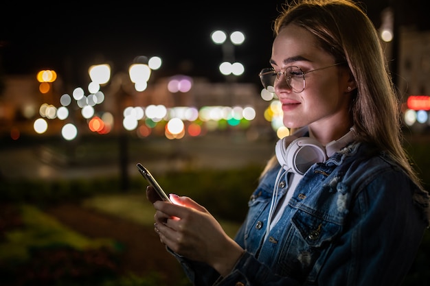 Vrolijke mooie jonge vrouw in koele bril en trendy slijtage die op metropoolstraat lopen met nachtlichten die van online playlistliederen in oortelefoons genieten en sms lezen met goed nieuws op smartphone