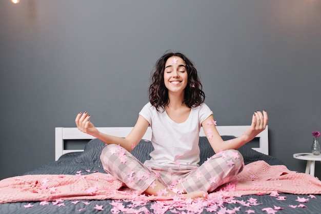 Vrolijke mooie jonge vrouw in pyjama's met krullend donkerbruin haar mediteren op bed in roze tinsels. gelukkig model lachend met gesloten ogen, ware emoties uitend, genietend van thuiscomfort