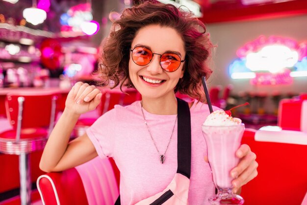 Vrolijke mooie stijlvolle lachende vrouw in kleurrijke retro vintage Amerikaanse jaren 50 café zittend aan tafel milkshake cocktail drinken met roze zonnebril plezier