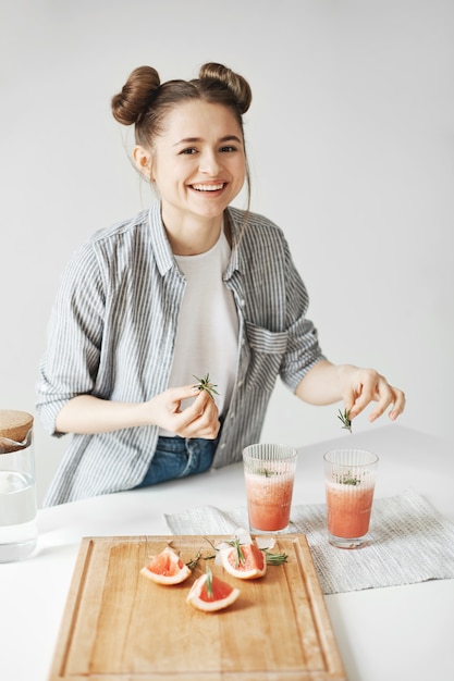 Vrolijke mooie vrouw die lacht versieren grapefruit detox smoothie met rozemarijn over witte muur. gezonde voeding.