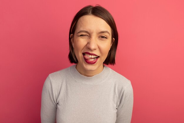 Vrolijke mooie vrouw knippert in het oog en steekt tong uit die op roze muur wordt geïsoleerd