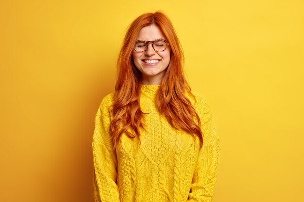 Vrolijke mooie vrouw met rood haar sluit ogen staat tevreden gekleed in casual trui draagt optische bril herinnert somethig zeer aangenaam.