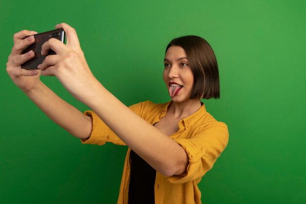 Vrolijke mooie vrouw steekt tong uit die selfie neemt die op groene muur wordt geïsoleerd