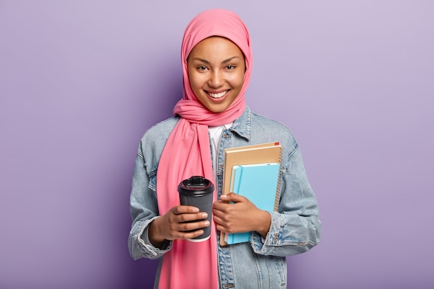 Vrolijke moslimvrouw in roze hijab, denim jas, draagt portemonnee