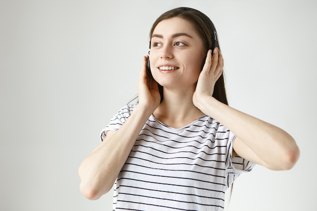 Vrolijke onbezorgde jonge donkerharige vrouw van in de twintig die binnenshuis ontspannen in vrijetijdskleding en draadloze hoofdtelefoons