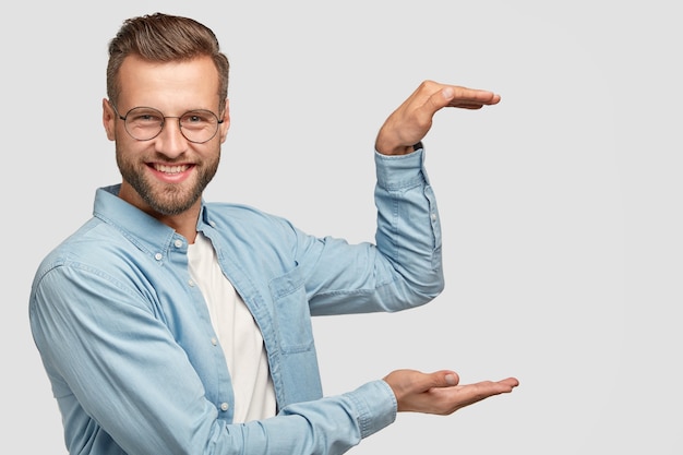 Gratis foto vrolijke ongeschoren man staat zijwaarts, houdt achtergrond vast, gebaart met beide handen alsof hij iets draagt, toont de hoogte van het ding