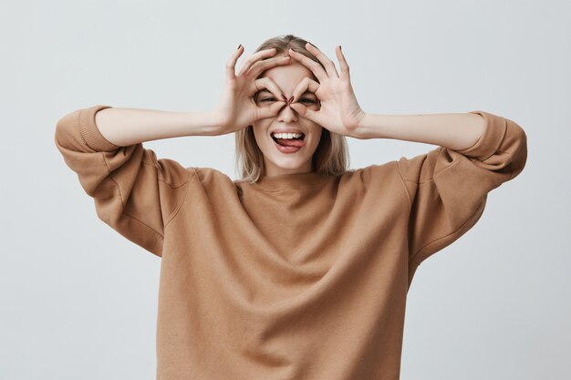 Vrolijke opgewonden vrouw met blond lang steil haar die ok-gebaren met beide handen laat zien, doet alsof ze een bril draagt en breed lacht, genietend van haar zorgeloos gelukkig leven