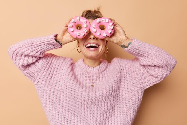 Vrolijke optimistische jonge vrouw bedekt ogen met heerlijke zoete donuts glimlacht in grote lijnen heeft plezier eet junkfood met veel calorieën draagt casual gebreide trui geïsoleerd over beige achtergrond.