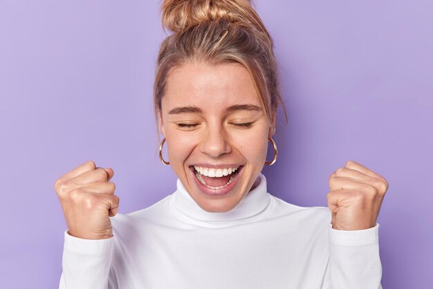 Vrolijke optimistische vrouw met haar knot houdt ogen gesloten glimlacht breed balt vuisten roept uit van vreugde viert goed nieuws draagt witte trui geïsoleerd over paarse achtergrond wint en verheugt zich