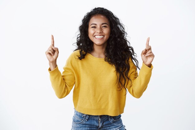 Vrolijke optimsitische jonge vrouwelijke student met krullend donker kapsel, gele trui, vrolijk lachend en lachend, wijzende vingers omhoog, vrienden laten linken naar site of kopieer ruimte, witte muur