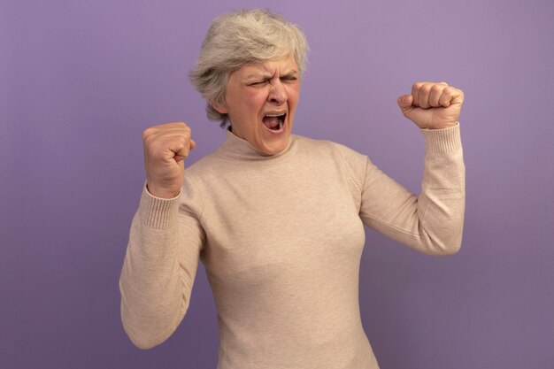 Vrolijke oude vrouw met een romige coltrui die een ja-gebaar doet met gesloten ogen geïsoleerd op een paarse muur