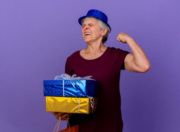 Vrolijke oudere vrouw met feestmuts houdt geschenkdoos en gespannen biceps geïsoleerd op paarse muur met kopie ruimte