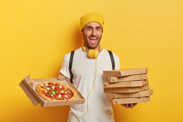 Vrolijke pizzabezorger staat met kartonnen dozen, wacht op klant, draagt gele hoed en wit t-shirt, luistert naar muziek tijdens het vervoer van fastfood
