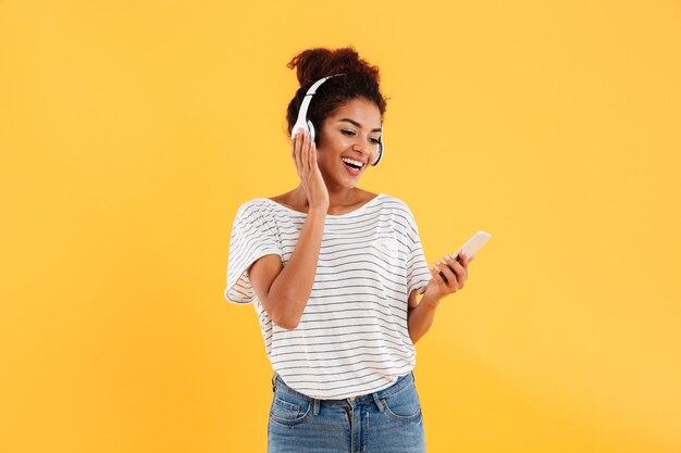 Vrolijke positieve jonge vrouw in hoofdtelefoons die geïsoleerde telefoon met behulp van