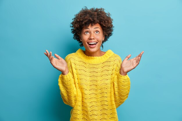 Vrolijke positieve vrouw met verwonderde blije uitdrukking kijkt naar boven en houdt de handpalmen omhoog glimlachen vrolijk gekleed in gebreide gele trui geïsoleerd op blauwe muur kan niet geloven dat droom uitkwam