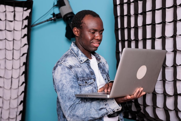 Vrolijke productiedirecteur met dslr-camera en handcomputer op blauwe achtergrond. glimlachende fotograaf die op laptop werkt terwijl hij in een studio staat die is uitgerust met professionele verlichting en softboxen.