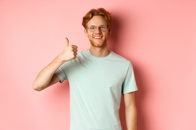 Vrolijke roodharige man in bril en t-shirt, duimen opdagen met een tevreden gezicht, positieve reactie tonen, goedkeuren en met je eens zijn, staande over roze achtergrond.