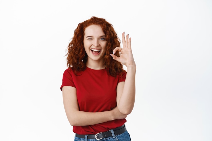 Gratis Foto vrolijke roodharige vrouw met krullend haar met ok-teken, knipogen en glimlachen, zeg ja, moedig aan om iets te kopen, maak een goed gebaar, keur en prijs een product van goede kwaliteit