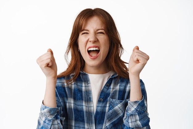 Vrolijke roodharige vrouw schreeuwen van vreugde en geluk, winnen, succes en overwinning vieren, doel bereiken, handen schudden en ja schreeuwen, staande op witte achtergrond