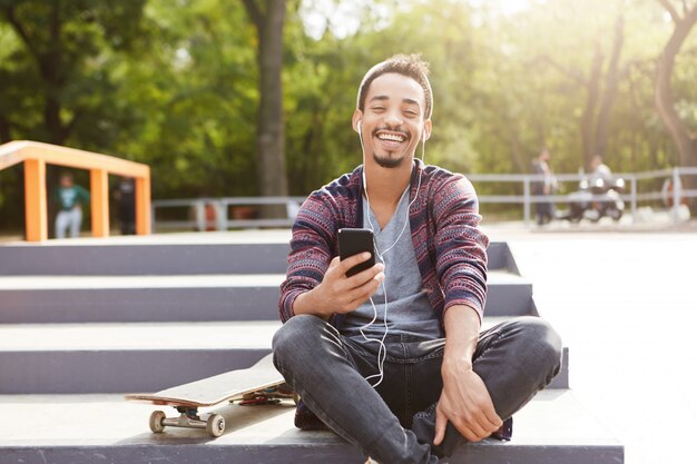 Vrolijke rustgevende gemengd ras tiener zit buiten op de grond, maakt gebruik van mobiele telefoon als melodieën met koptelefoon luistert