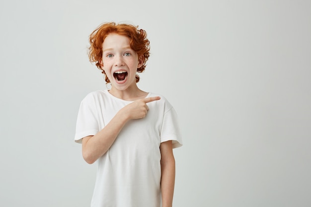 Vrolijke schattige kleine jongen met krullend gember haar en sproeten gelukkig lachend en opzij wijzen