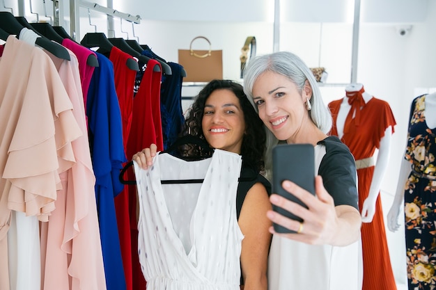 Vrolijke schattige vriendinnen genieten van winkelen in boetiek samen, jurk houden, plezier hebben en selfie op mobiele telefoon te nemen. Consumentisme of winkelconcept