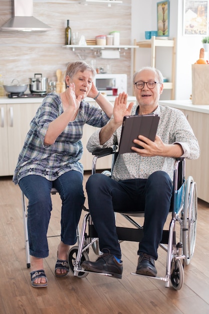 Vrolijke senior vrouw zwaaien op videoconferentie in de keuken. Gehandicapte senior man in rolstoel en zijn vrouw hebben een videoconferentie op tablet-pc in de keuken. Verlamde oude man en zijn vrouw hebben...