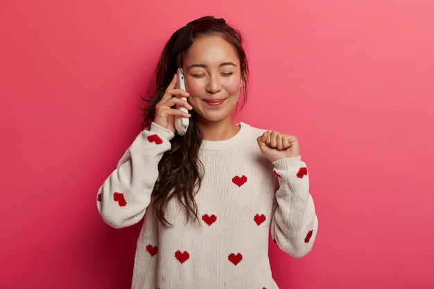 Vrolijke tienermeisje heeft telefoongesprek via mobiele telefoon, verheugt zich goed nieuws, gebalde vuist verhoogt, geniet van gelukkige communicatie, ogen sluit