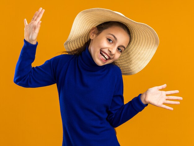 Vrolijke tienermeisje met een strandhoed die naar de voorkant kijkt en lege handen toont die op een oranje muur zijn geïsoleerd