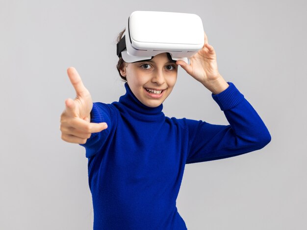 Vrolijke tienermeisje met een vr-headset die het opheft en naar de voorkant wijst, geïsoleerd op een witte muur