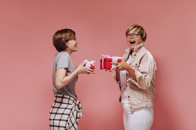 Vrolijke twee vrouwen met kort modern kapsel in stijlvolle kleding en glazen met rode geschenkdozen en vreugde op roze achtergrond.