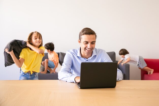Vrolijke vader chatten via laptop en kinderen spelen met kussens in de buurt van hem. Kaukasische vader thuis werken tijdens schoolvakanties. Familie en digitale technologie concept