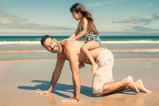 Vrolijke vader gaat op handen en knieën op het strand, met meisje op zijn rug