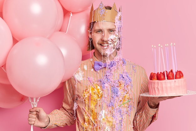 Vrolijke verjaardag man met blije uitdrukking draagt papieren kroon vuile feestelijke kleding houdt cake en ballonnen vormt op feestje tegen roze muur viert verjaardag of krijgt nieuwe positie