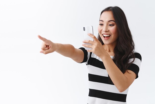 Vrolijke vermaakte enthousiaste jonge vrolijke aziatische vrouw in casual outfit video opnemen als het controleren van iets geweldigs wijzende vinger zijwaarts fotograferen verlicht concert witte achtergrond