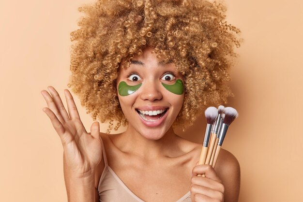 Vrolijke verraste vrouw met krullend haar houdt palm omhoog staart afgeluisterde ogen past groene patches toe om de huid te verfrissen houdt cosmetische borstels geïsoleerd over bruine achtergrond Gezichtsbehandelingen concept