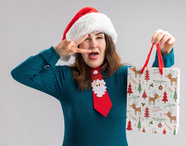 Vrolijke volwassen blanke vrouw met kerstmuts en stropdas santa stucks tong en houdt papier geschenkdoos geïsoleerd op een witte achtergrond met kopie ruimte