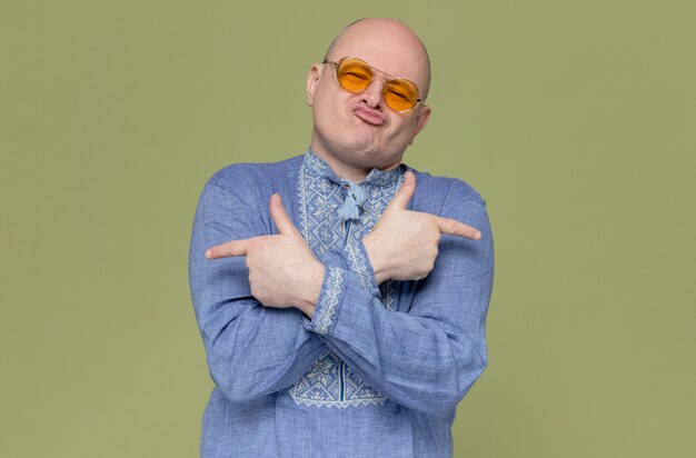 Vrolijke volwassen Slavische man in blauw shirt met een zonnebril die naar de zijkanten wijst
