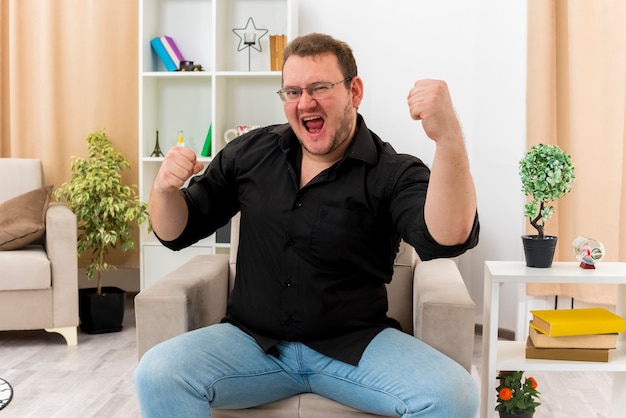 Vrolijke volwassen Slavische man in optische bril zit op fauteuil houden vuisten kijken camera in de woonkamer