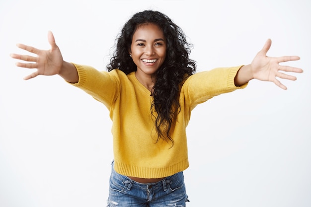 Gratis foto vrolijke vriendelijk ogende mooie vrouw in gele trui, handen gespreid in een warm welkom, vrolijk glimlachend wil knuffelen, groet vriend, knuffelen