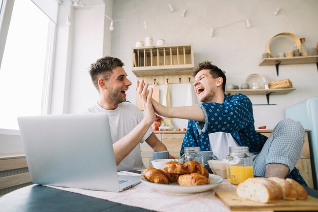 Vrolijke vrienden die handen slaan die voor lijst met ontbijt en laptop zitten